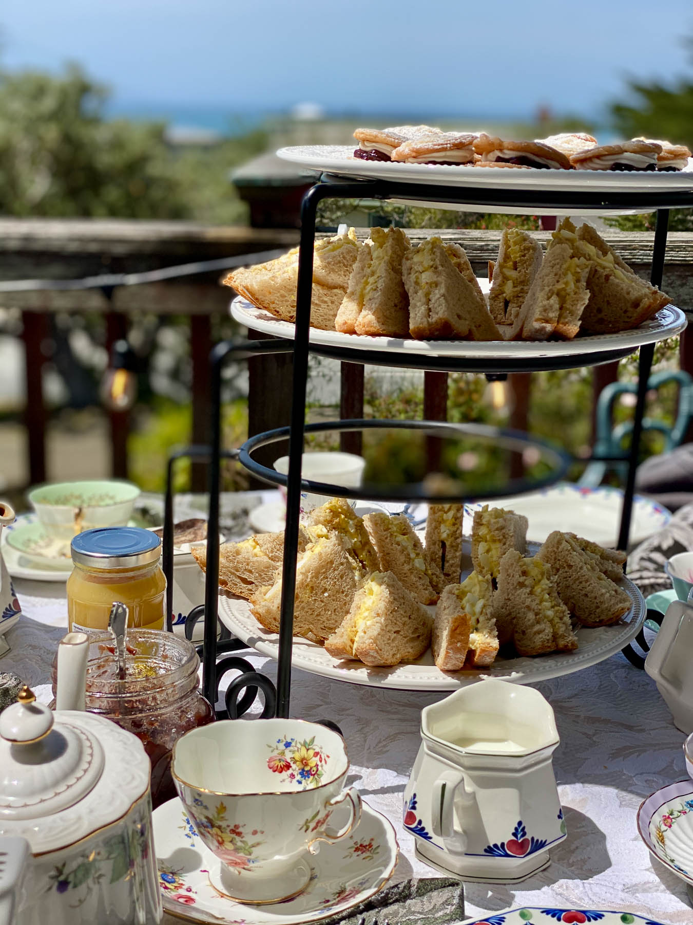 A three-tiered stand filled with assorted tea sandwiches on a table set with floral china teacups, a teapot, and jam for an outdoor afternoon tea party.