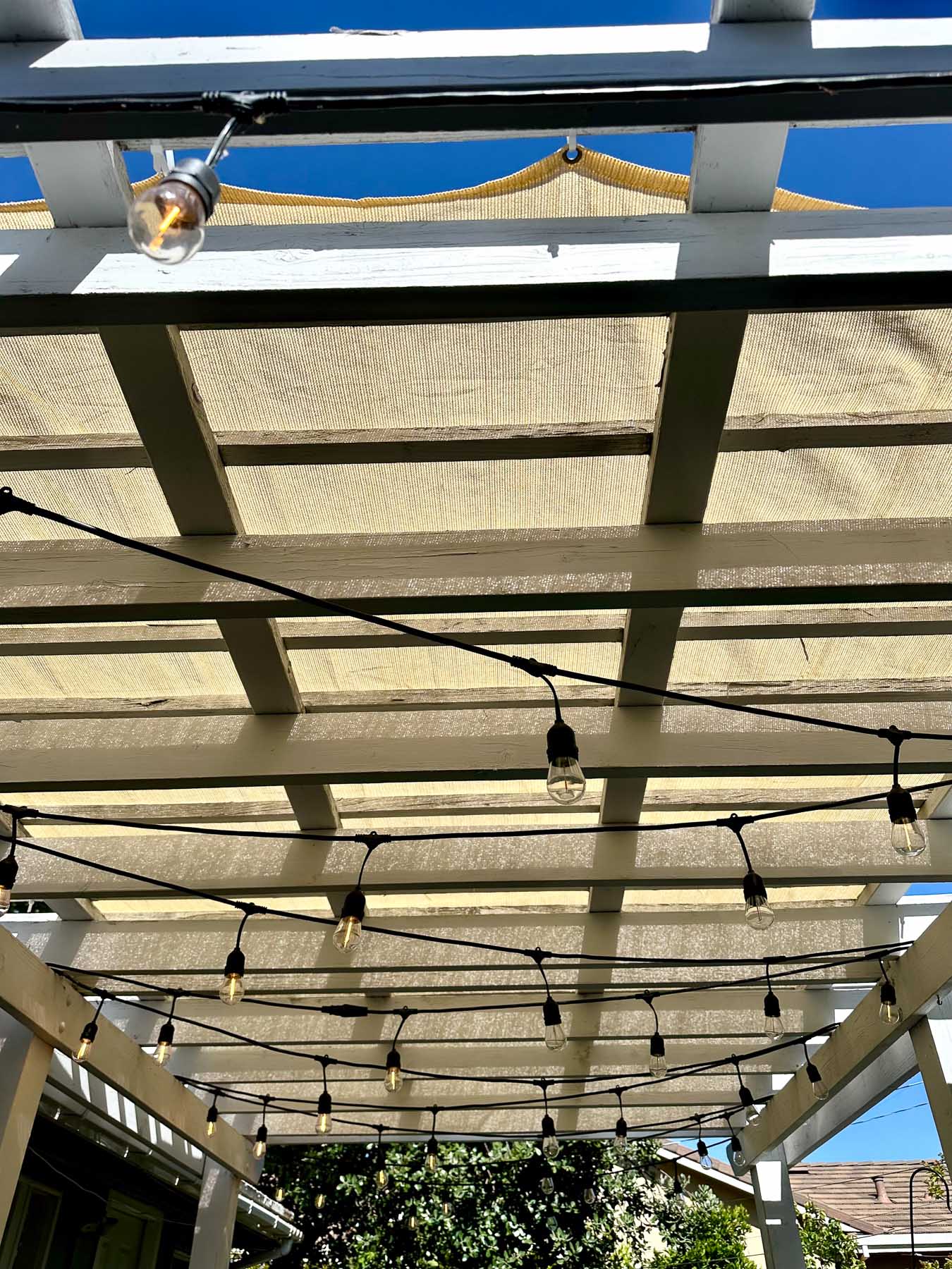 Looking up at a white pergola fitted with a tan pergola shade and string lights, providing comfortable backyard shade.