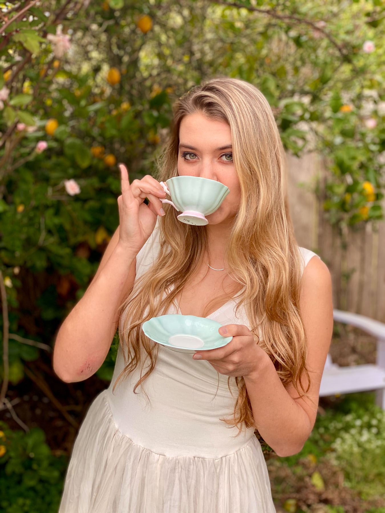 A young woman in a white dress sipping from a delicate light green teacup while standing in a lush backyard garden.