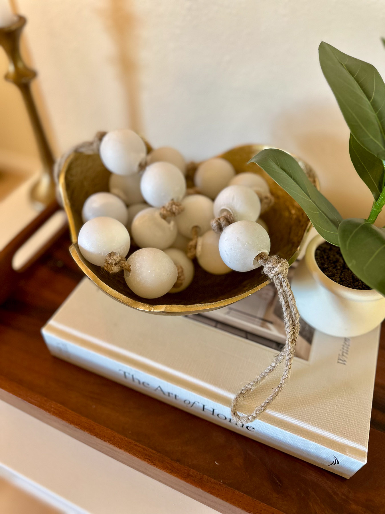 A textured gold bowl holding a garland of large, white seaglass-style beads strung on jute rope, displayed next to a green houseplant.