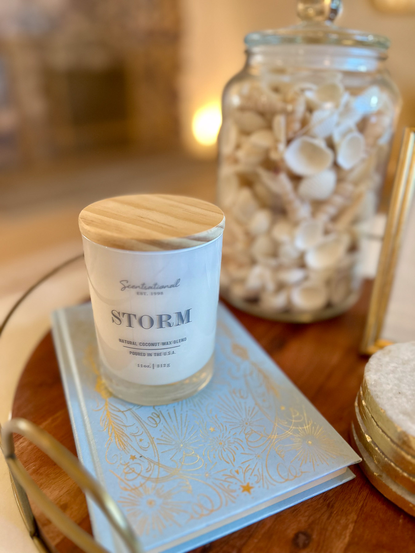 A coastal vignette featuring a coconut wax blend candle with a wooden lid resting on a light blue book with gold details, next to a jar of seashells.