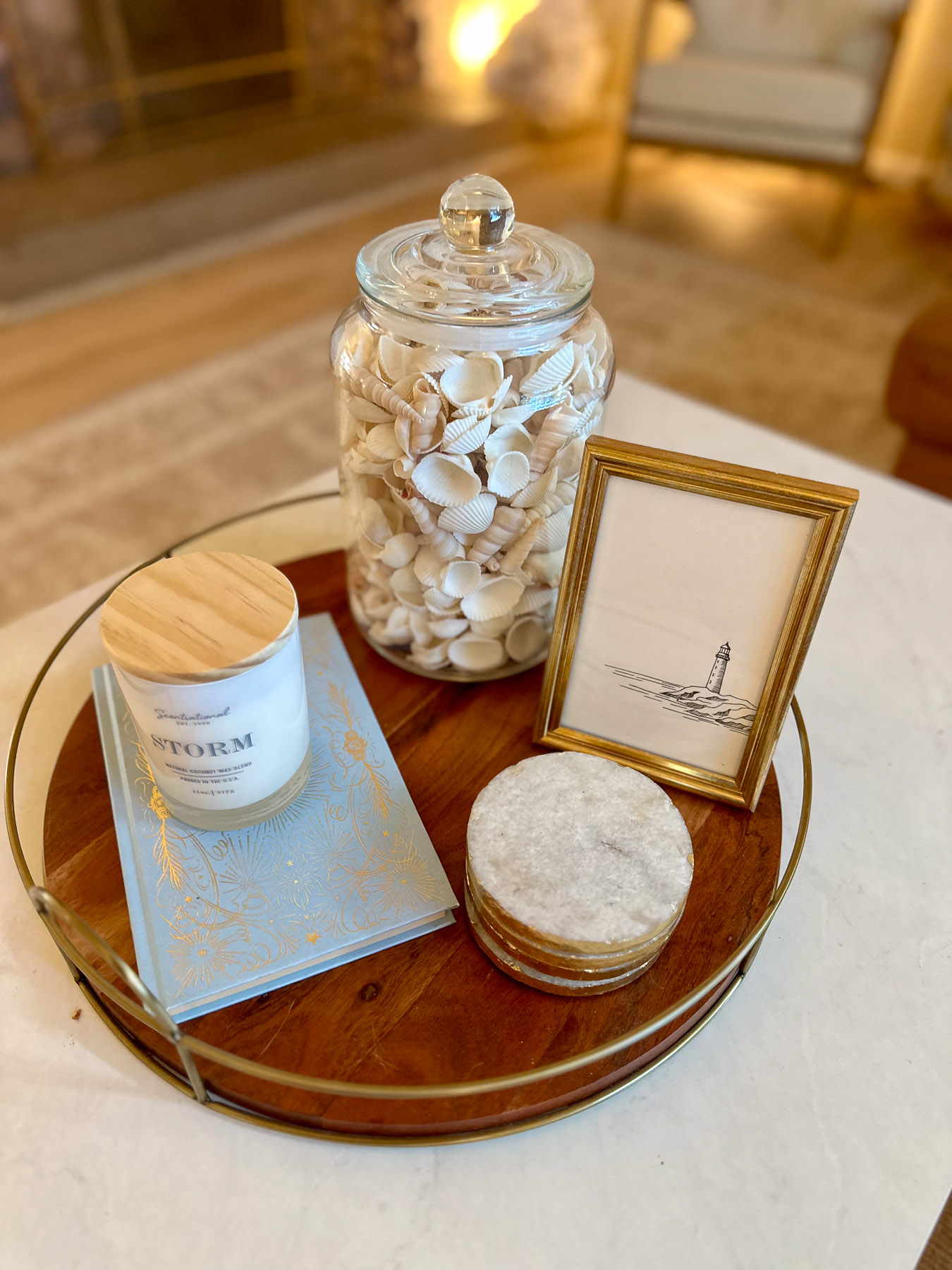 A wooden tray holding a large glass jar filled with seashells, a blue book, a white candle, and a framed lighthouse print.
