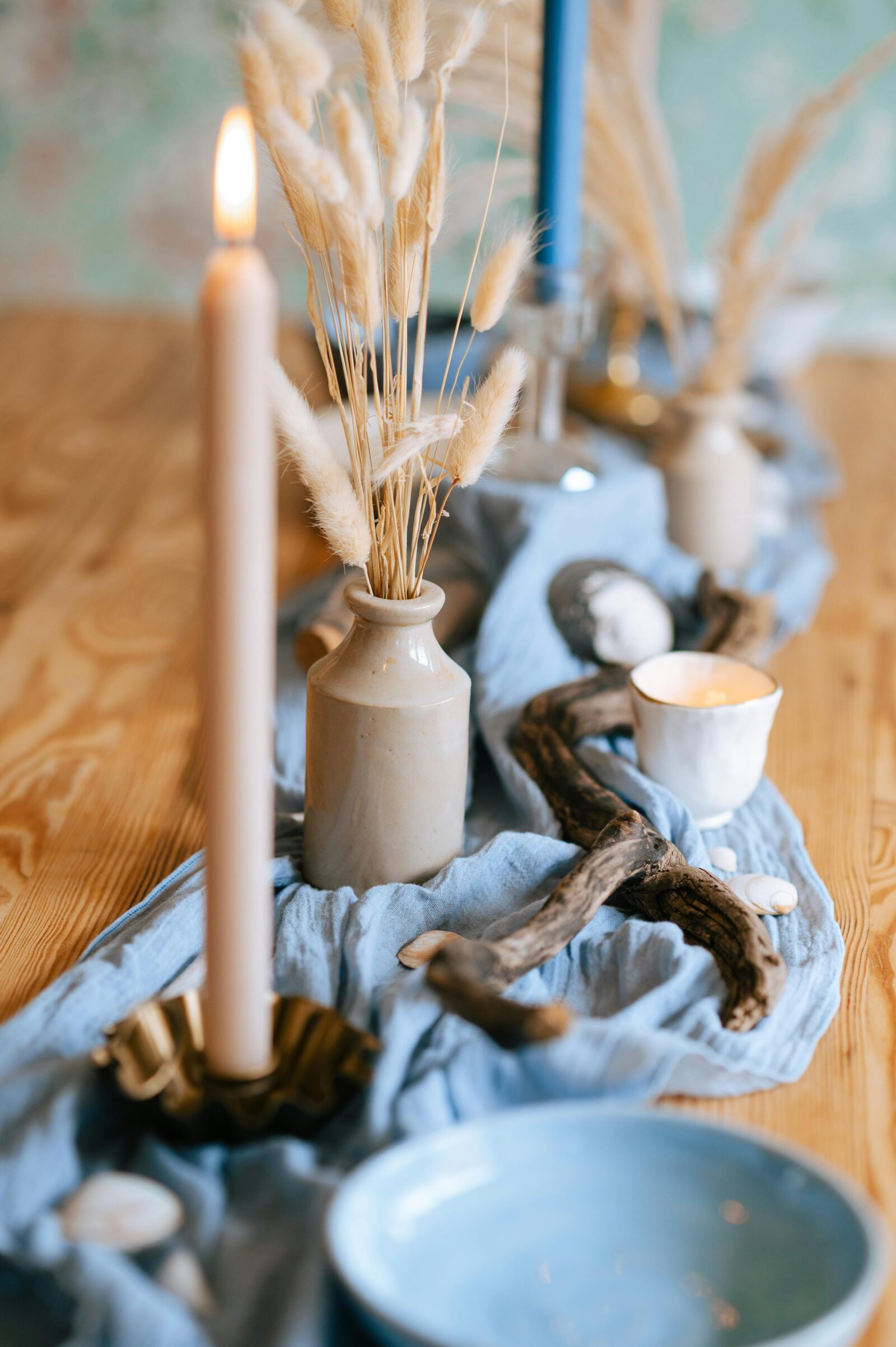 A coastal centerpiece featuring dried bunny tails in a ceramic vase, driftwood, and a taper candle on a blue table runner.