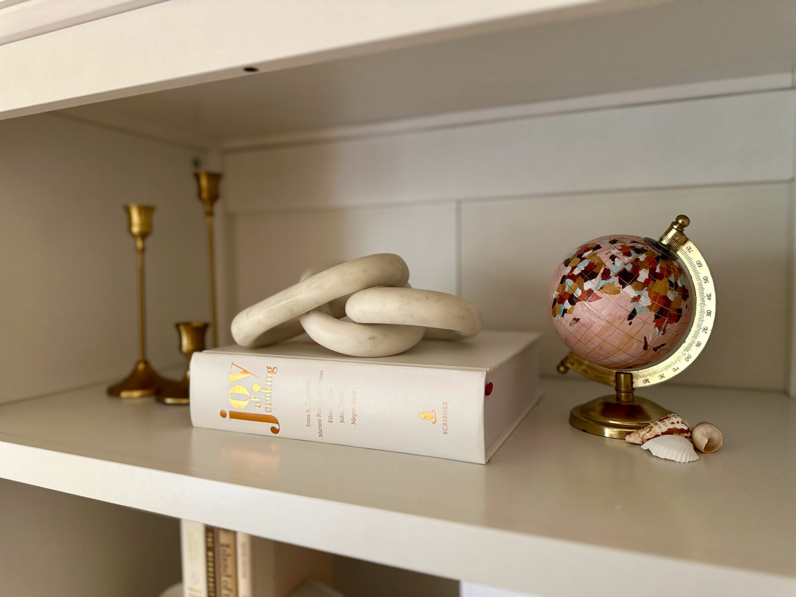 A minimalist coastal vignette on a white bookshelf featuring a white marble chain link resting on a white Joy of Cooking book, next to a decorative globe.