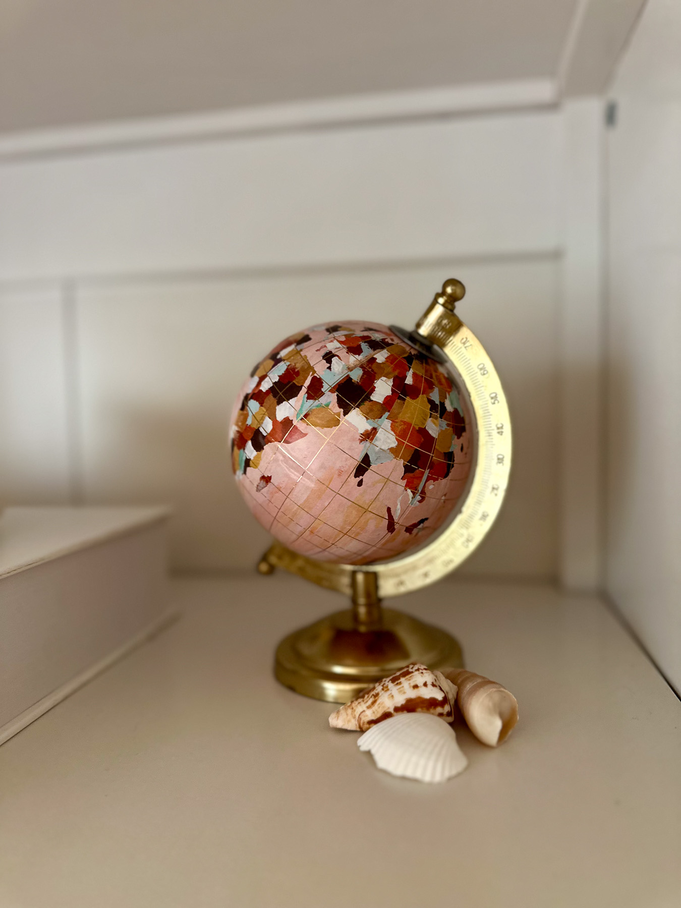 Close-up of a small pink and gold decorative globe resting on a white shelf next to scattered seashells.