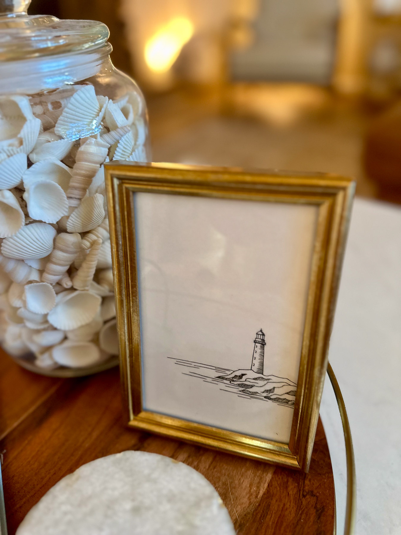A minimalist black and white framed sketch of a lighthouse styled next to a jar of white seashells on a wooden tray.