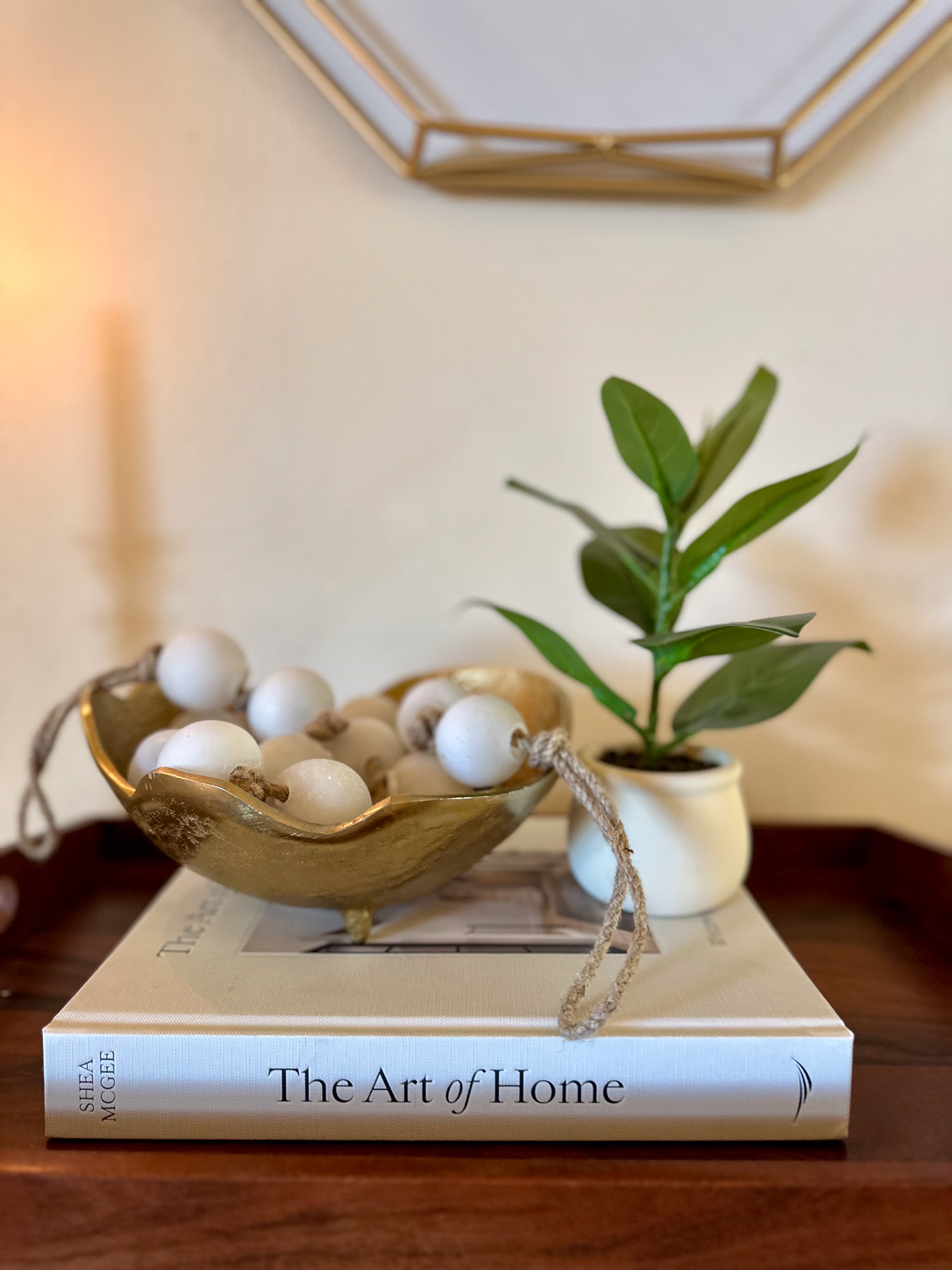 A gold bowl filled with white decorative beads and a small potted green houseplant resting on a coffee table book titled The Art of Home.