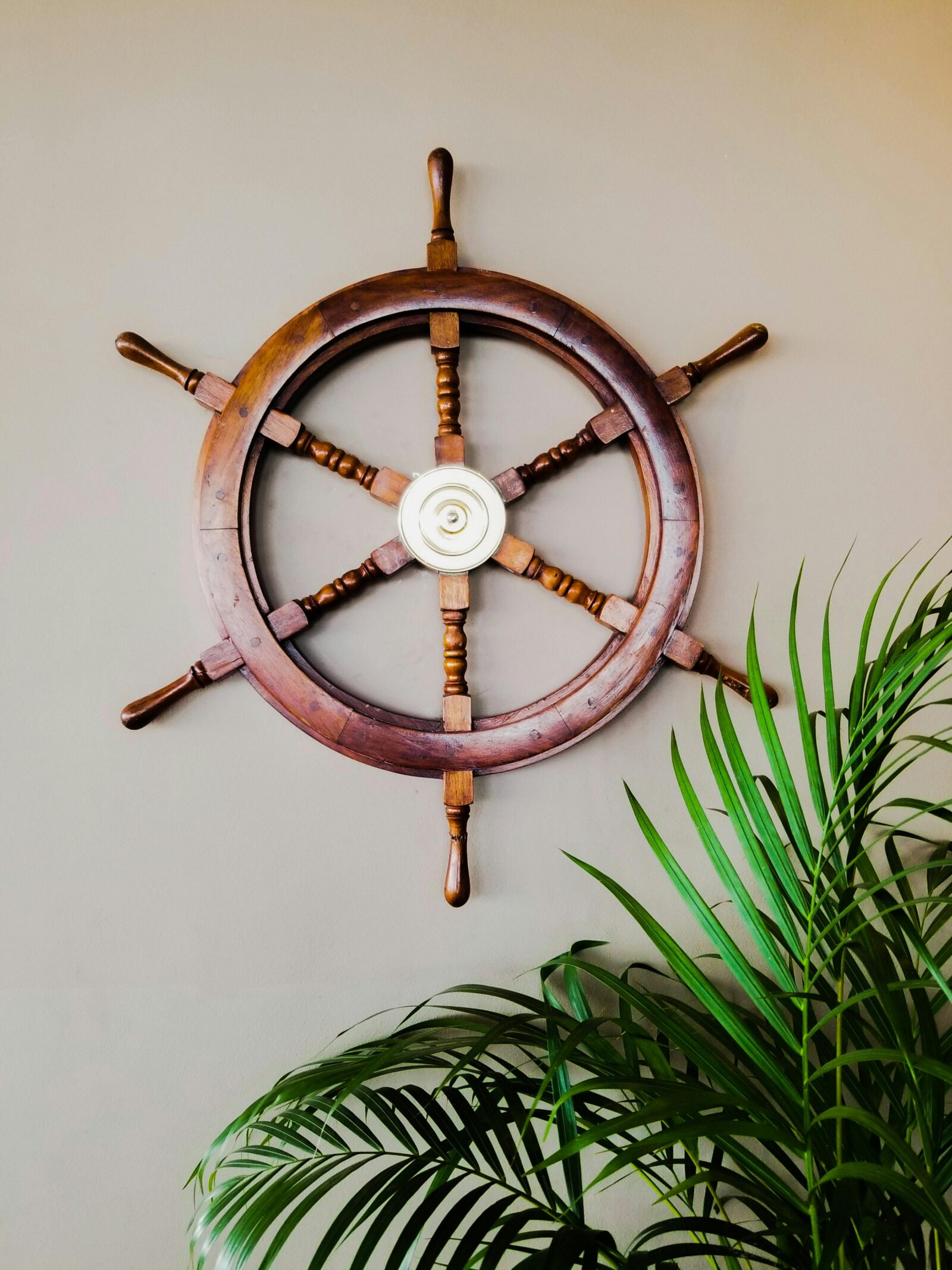 A classic wooden ship's steering wheel mounted on a light wall above lush green palm leaves.